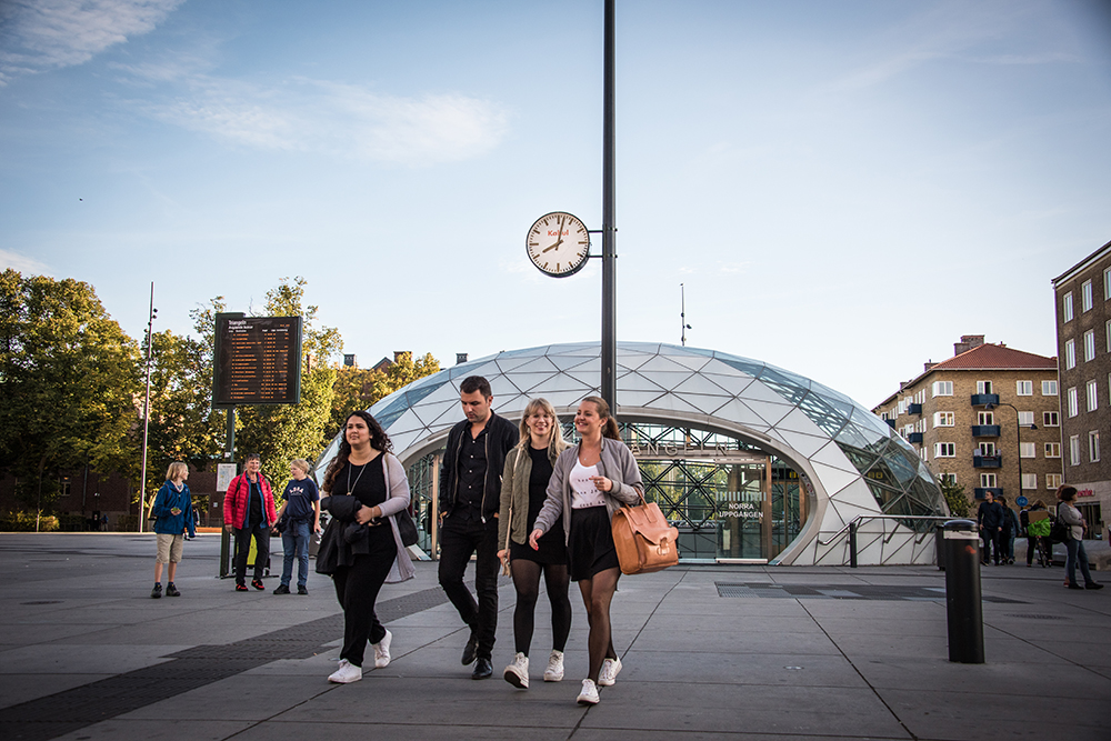 En grupp människor lämnar den glasöverbyggda utgången från station Triangeln. Ovanför deras huvuden ser man en vanlig offentlig klocka med texten "Kabul" i rött. Jens Haaning, Kabul Time.
