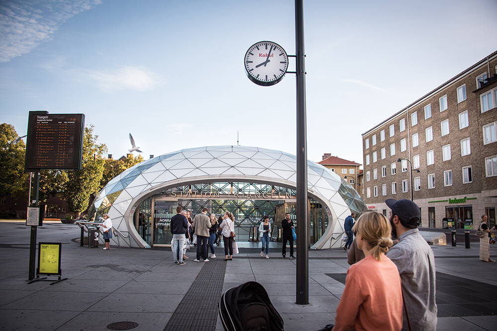 Folk går in och ut i den glasöverbyggda entrén till station Triangeln. Ovanför deras huvuden ser man en vanlig offentlig klocka med texten "Kabul" i rött. Jens Haaning, Kabul Time.