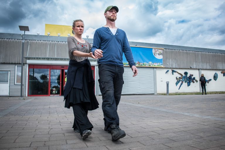 En kvinna med öppna ögon leder en man vars ögon är slutna, genom att hålla honom vid ena armbågen och underarmen. De går bredvid varandra på en stenlagd plats. Myriam Lefkowitz, Walk, hands, eyes (Gamlegården)