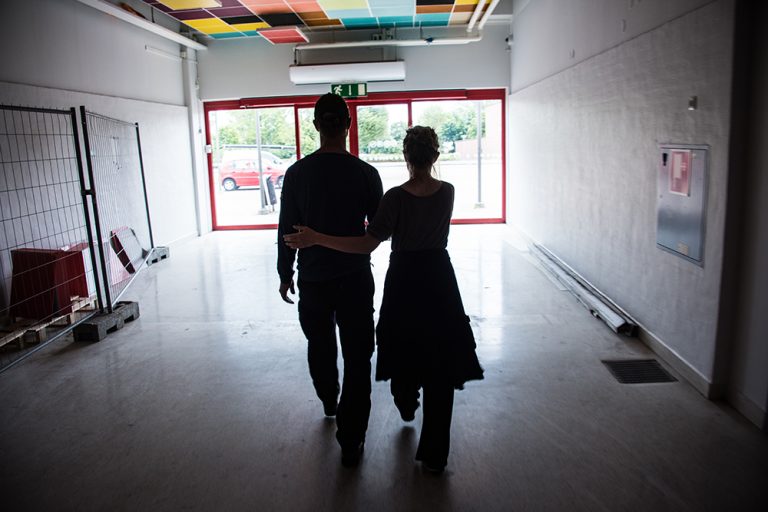 En man och en kvinna går bort från kameran mot en utgång. Kvinnan leder mannen genom att hålla med ena handen i hans vänstra armbåge och med den andra i hans högra underarm. Myriam Lefkowitz, Walk, hands, eyes (Gamlegården)