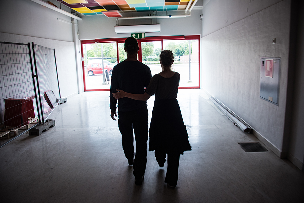 En man och en kvinna går bort från kameran mot en utgång. Kvinnan leder mannen genom att hålla med ena handen i hans vänstra armbåge och med den andra i hans högra underarm. Myriam Lefkowitz, Walk, hands, eyes (Gamlegården)