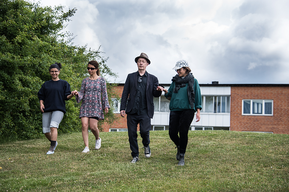 Två kvinnor med öppna ögon leder en man respektive en kvinna vars ögon är slutna nerför en kulle. Bakom dem ett bostadshus. Myriam Lefkowitz, Walk, hands, eyes (Gamlegården)