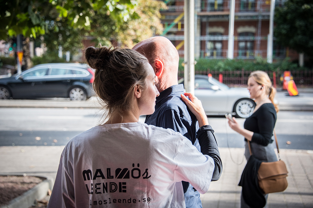 En kvinna i vit t-tröja står med ryggen mot kameran. Hon guidar en man som står framför henne, halvt dold för kameran. Myriam Lefkowitz, Walk, Hands, Eyes (Malmö)
