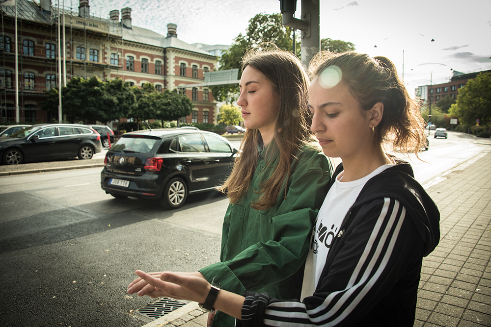 En kvinna med slutna ögon leds vid handen längs trottoaren av en annan kvinna. Bredvid dem kör bilar. Myriam Lefkowitz, Walk, Hands, Eyes (Malmö)