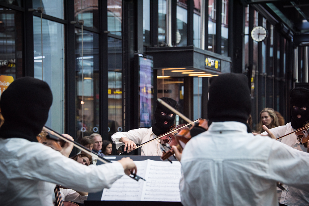 Flera violinister i vita skjortor och svarta ansiktsmasker som endast har hål för ögon och mun. Deras blickar möts över notstället medan de spelar. I bakgrunden publik. Sislej Xhafa, Again and again.