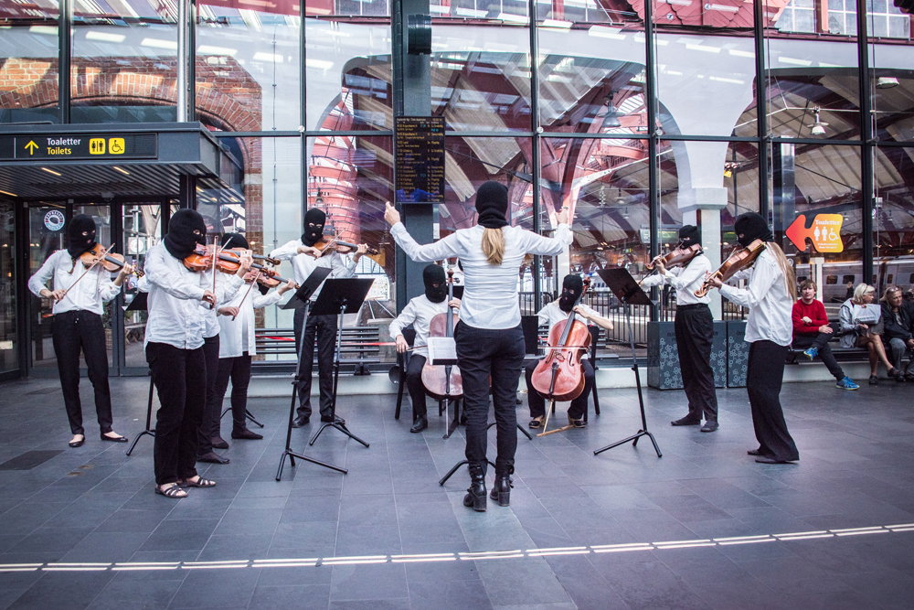 En spelande stråkensemble bestående av åtta musiker och en dirigent som har ryggen mot kameran. Alla har vita skjortor och svarta ansiktsmasker. Instrumenten är fiol, cello och viola. Sislej Xhafa, Again and again.