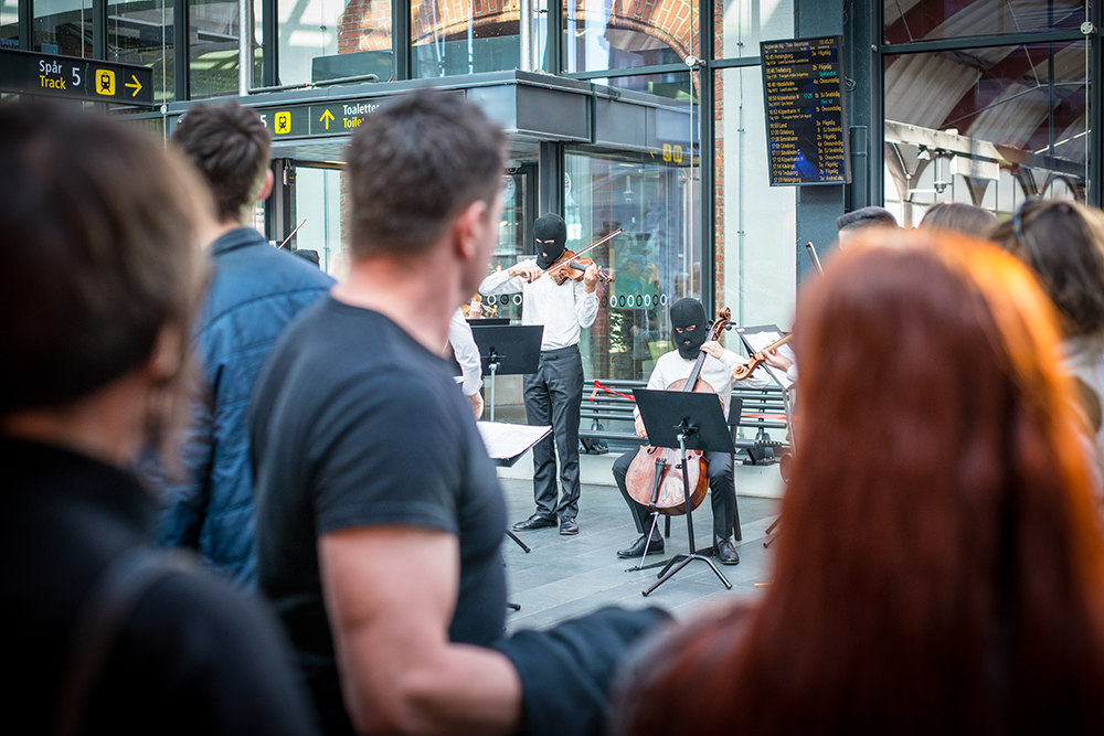 I förgrunden suddig publik som betraktar en spelande cellist och en violinist i svarta ansiktsmasker. I bakgrunden avgångstavlor för tågen. Sislej Xhafa, Again and again.