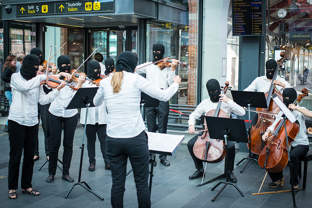 Dirigentens långa ljusa hår sticker ut under den svarta heltäckande ansiktsmasken. Framför henne sitter cellisterna och bredvid henne spelar violinisterna, alla med ansiktsmasker. Sislej Xhafa, Again and again.