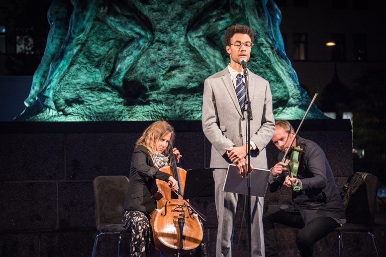 Konstnären Santiago Mostyn, iklädd slips och kostym, står på ett podium framför den upplysta statyn på Möllevångstorget. Bakom honom en cellist och en violinist. I bakgrunden torget och verket Mirakel av samme konstnär. Santiago Mostyn, The repetition och Mirakel.