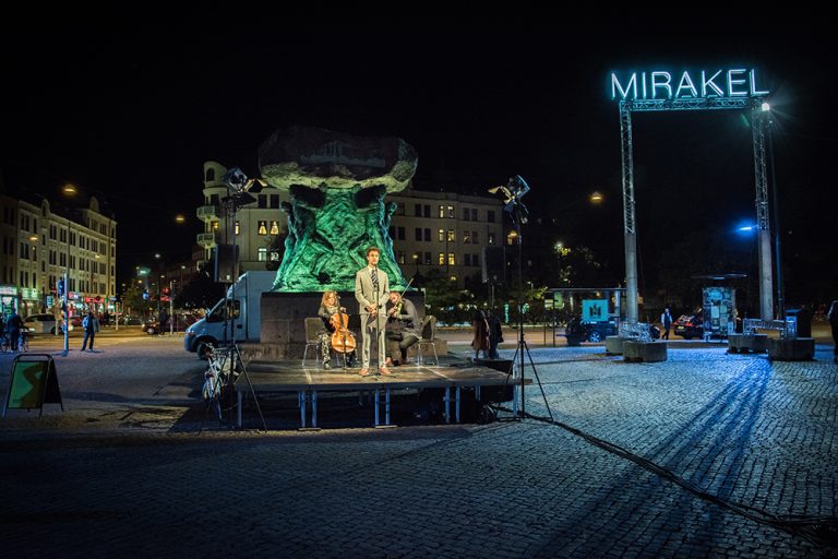 Konstnären Santiago Mostyn, iklädd slips och kostym, står på ett podium framför den upplysta statyn på Möllevångstorget. Bakom honom en cellist och en violinist. Santiago Mostyn, The repetition.