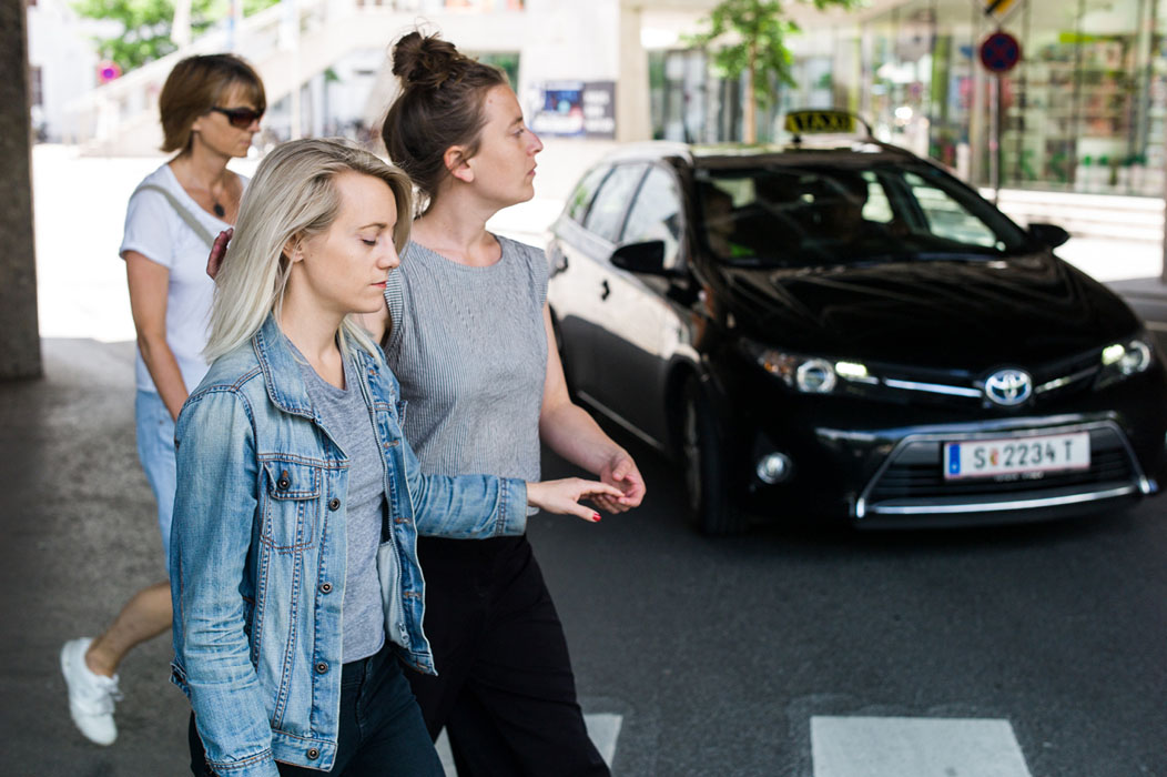 En kvinna med öppna ögon leder en kvinna vars ögon är slutna över ett övergångsställe, genom att hålla henne lätt i ena handens fingrar och i armbågen. Myriam Lefkowitz, Walk, hands, eyes (Gamlegården)