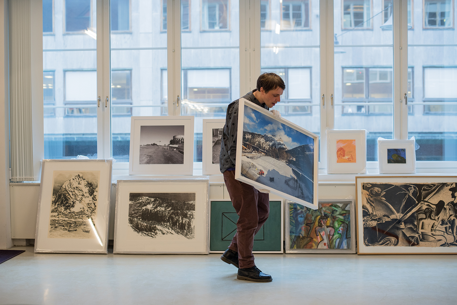 En man bär och tittar på ett konstverk. Tavlor står radade under fönster i bakgrunden.