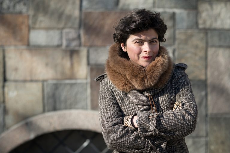 Kvinna står och tittar rakt in i kameran. Hon har en vinterkappa på sig. En stenmur i bakgrunden.