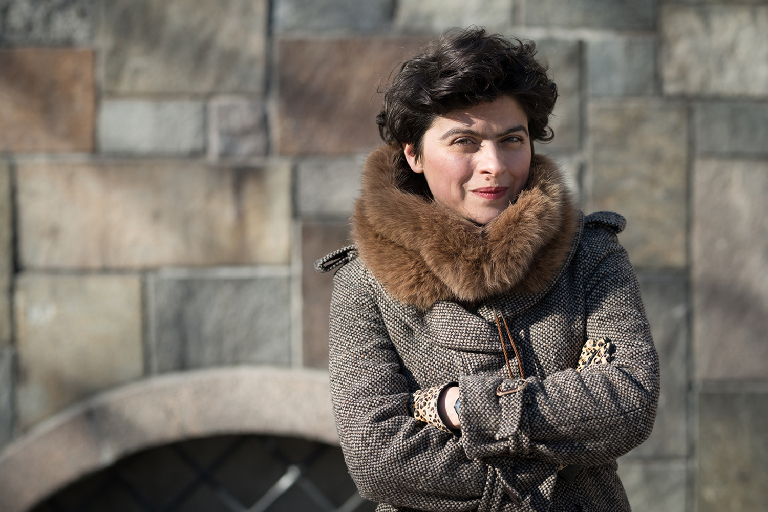 Kvinna står och tittar rakt in i kameran. Hon har en vinterkappa på sig. En stenmur i bakgrunden.