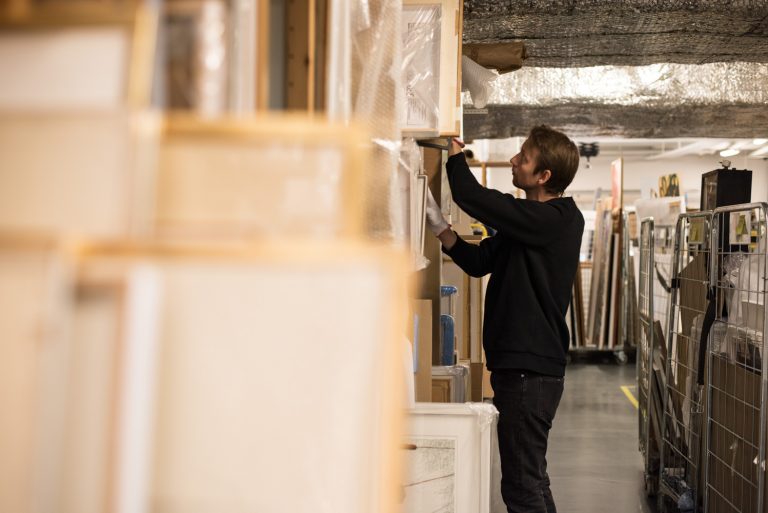 En man i gakgrunden arbetar med hyllor där det står tavlor.