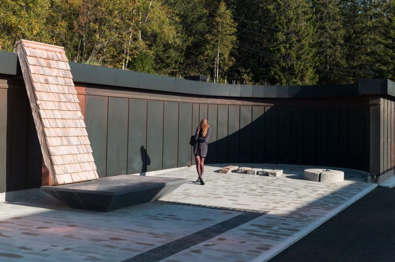 En tegelvägg står lutad mot ett hus. Den vilar på ett stenfundament. Det står en person bredvid skulpturen.