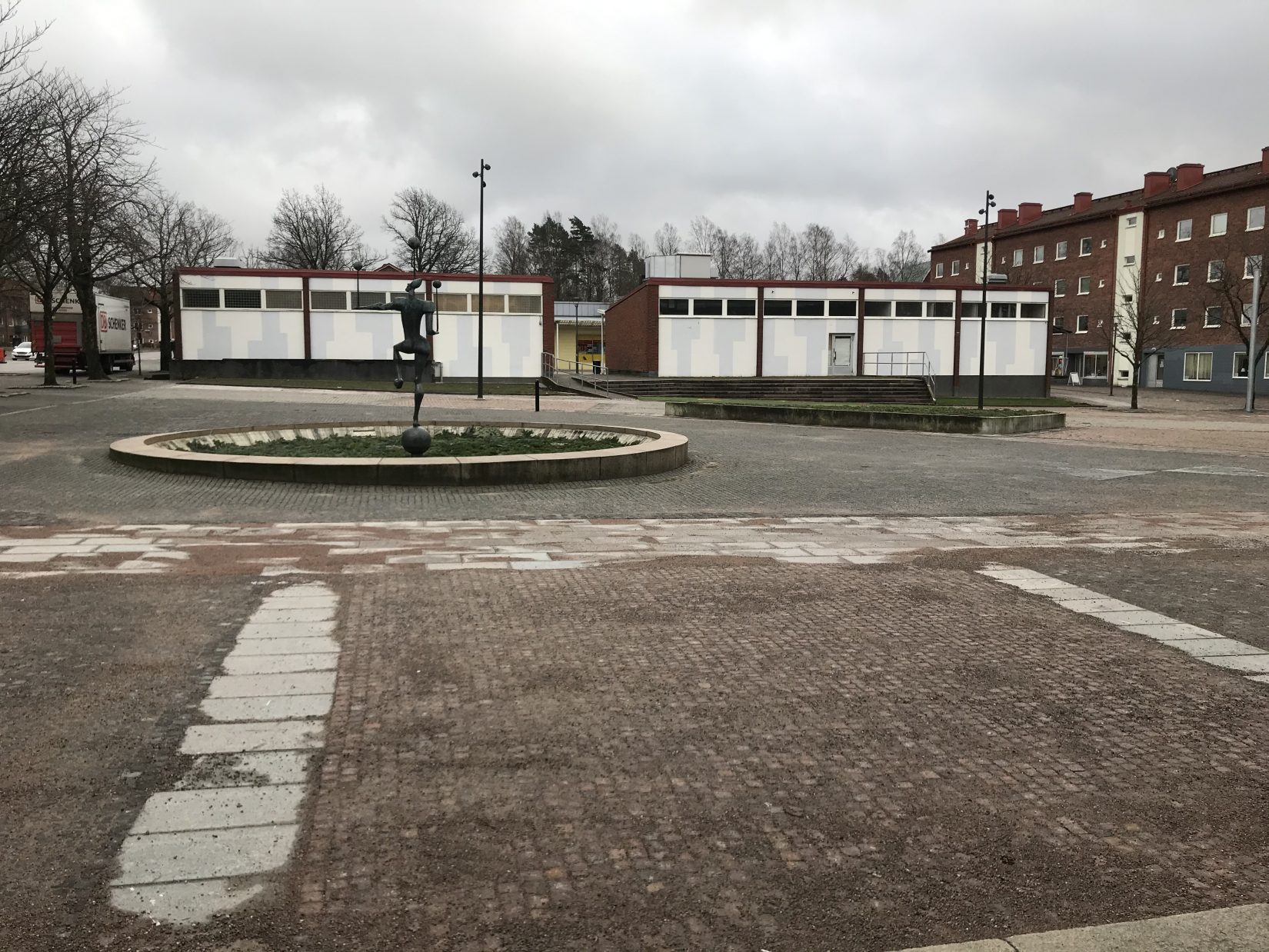 Ett torg med en staty i rundel med gräs. Två låga byggnader syns i bakgrunden.