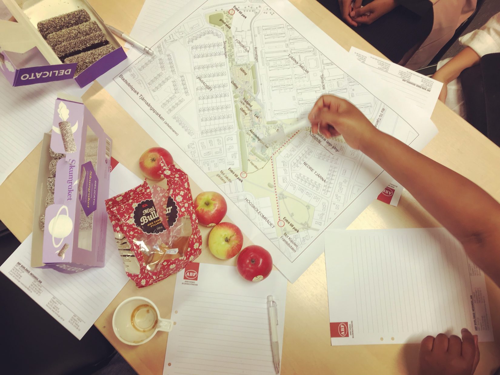 Bord sett rakt ovanifrån med kartor, papper, mat och händer.