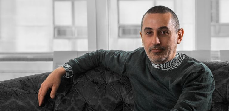 En man sitter lutad mot en soffa. Han tittar rakt in i kameran. Fönster i bakgrunden.