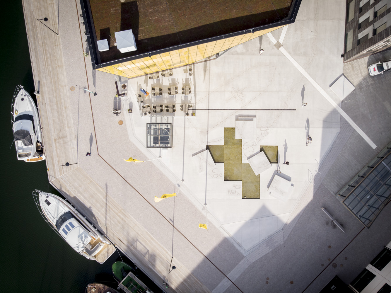 Bild på torg sett ovanifrån. Höger om torget syns båtar och vatten. Mitt på torget är stenplattor bortplockade och vatten syns. Konturer av ett skepp syns svagt i betongen-