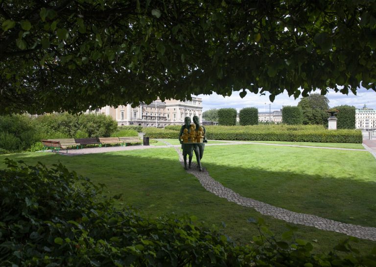 På en gräsmatta är en stenlagd stig. På stigen finns en skulptur av två unga kvinnor som bär fram byster på fem kvinnor.