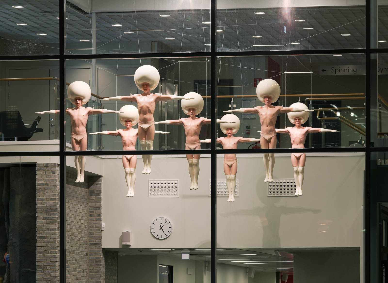 Seven sculptures in plastic by men dressed in white underwear wearing helmets. They are hanging in the air.