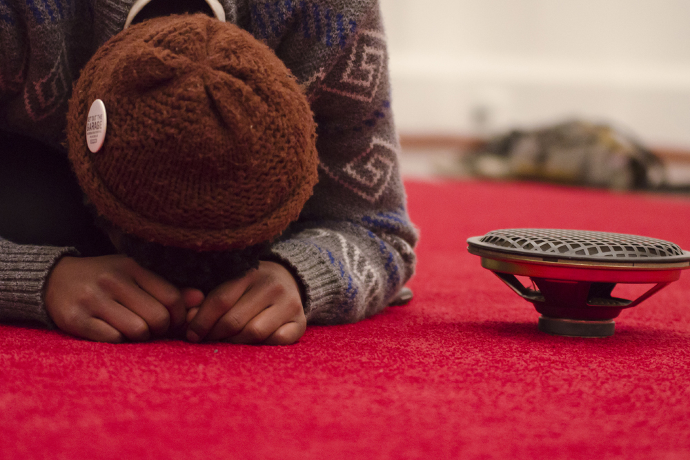 En person i kofta och mössa ligger framåtböjd på en röd matta bredvid en högtalare. James Webb, Prayer (Malmö).