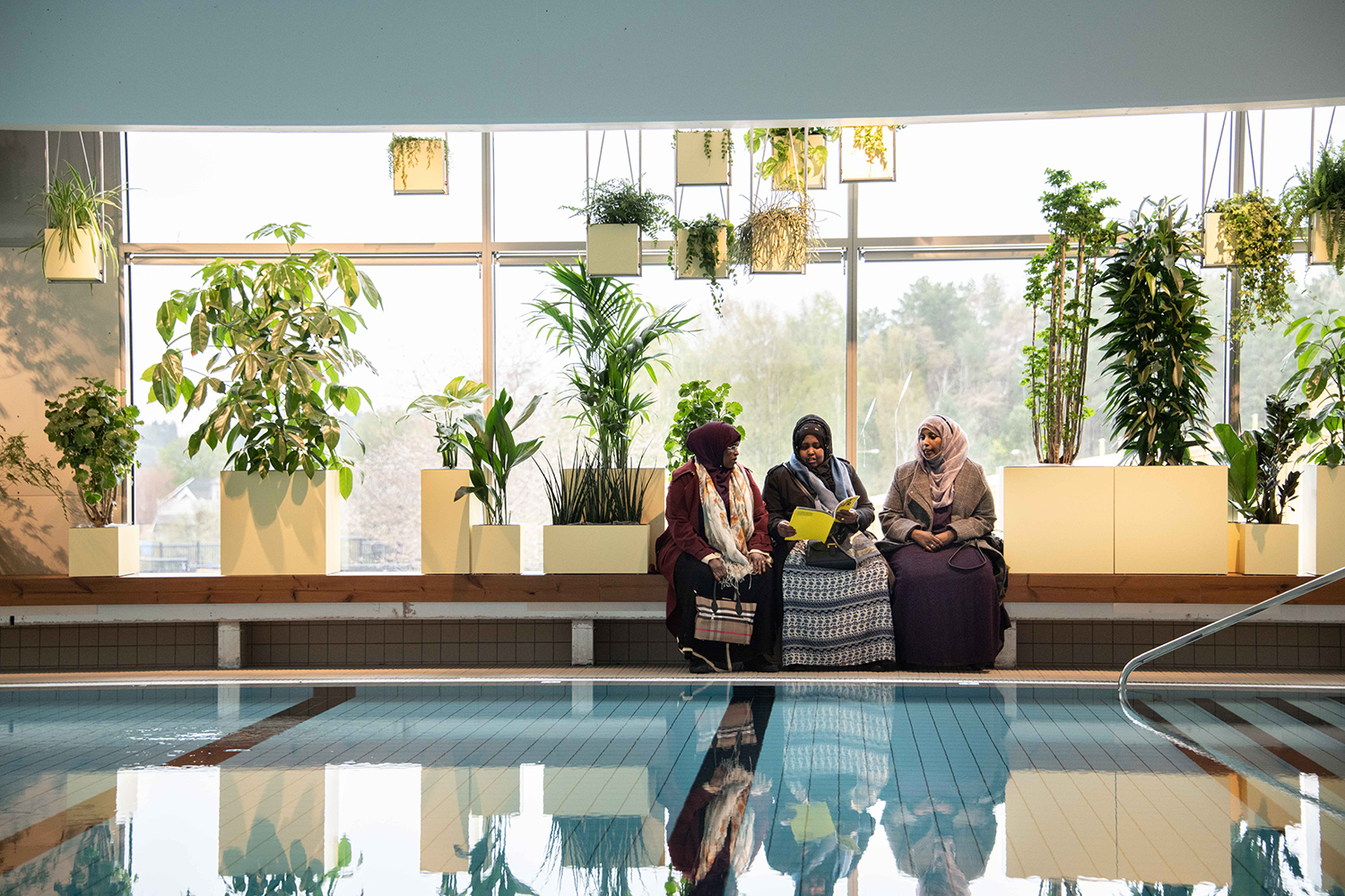 An indoor swimming pool with several plants in the background and three sitting women.