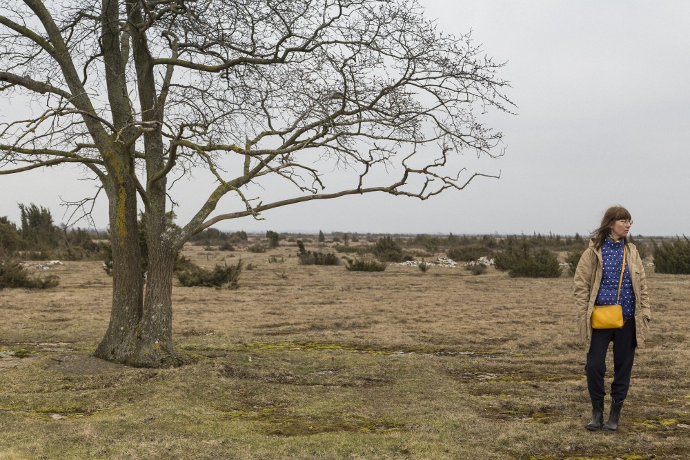 En kvinna står vid ett träd i Dröstorp Naturreservat på Öland. I bakgrunden gräs och låga buskar. Pia Sandström, Minnet Orden Marken..