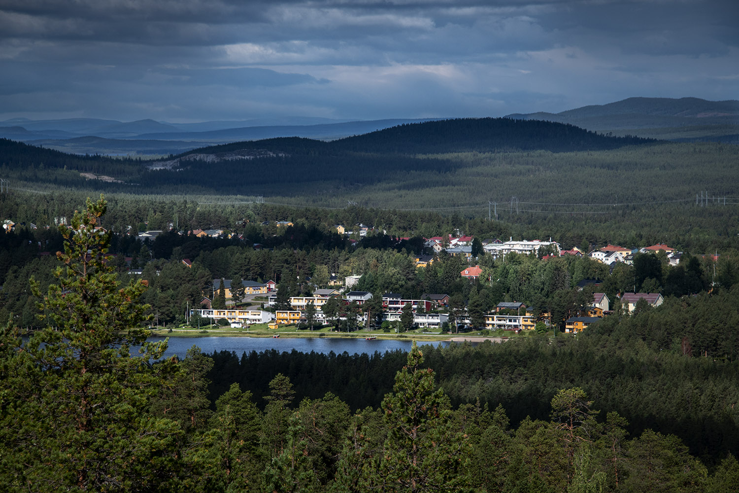 View over the Jokkmokk landscape with loots of pine trees, a lake, and a group of building.