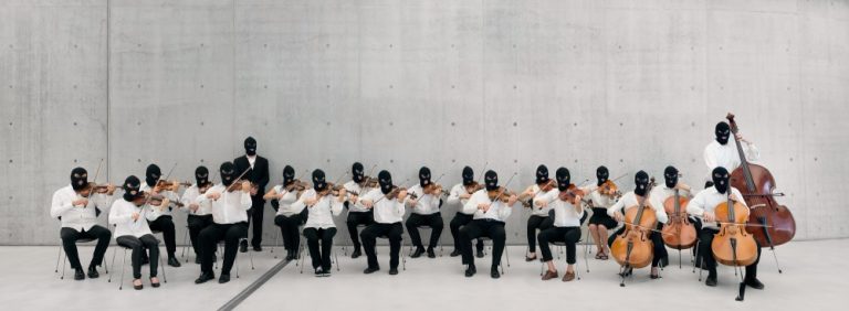 En tjugo personer stark stråkorkester med fioler, viola och cello. Alla musiker har vita skjortor och svarta rånarluvor. Orkestern sitter och spelar i ett kalt rum av betong. Sislej Xhafa, Again and Again