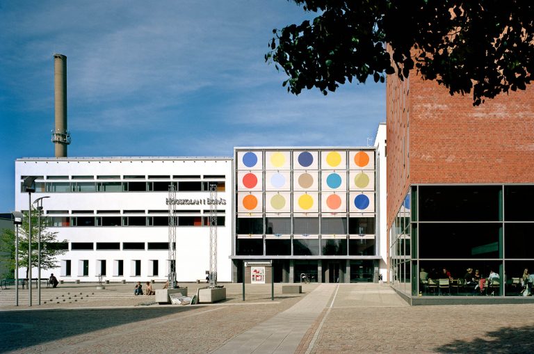 Högskolan i Borås utifrån. Ena flygeln av tegel, den andra vit. I mitten biblioteksbyggnaden med solavskärmningen halvvägs nerfälld och de olikfärgade prickarna väl synliga. Kontrapunkt, Eva Stephenson-Möller