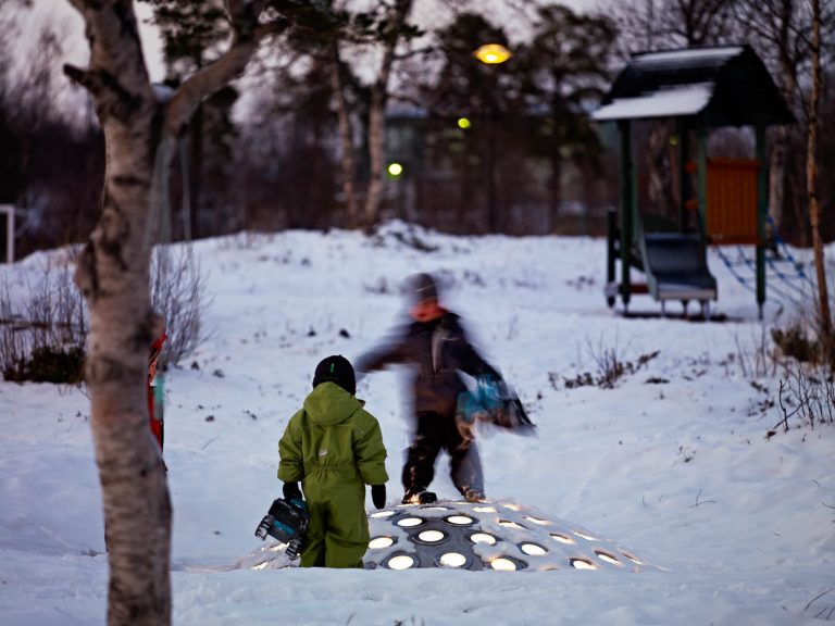 Två barn leker på stålkupolen som sticker upp ur snön. Lamporna lyser vita. I bakgrunden en lekplats. Markus Lantto, Termonoder, 2009