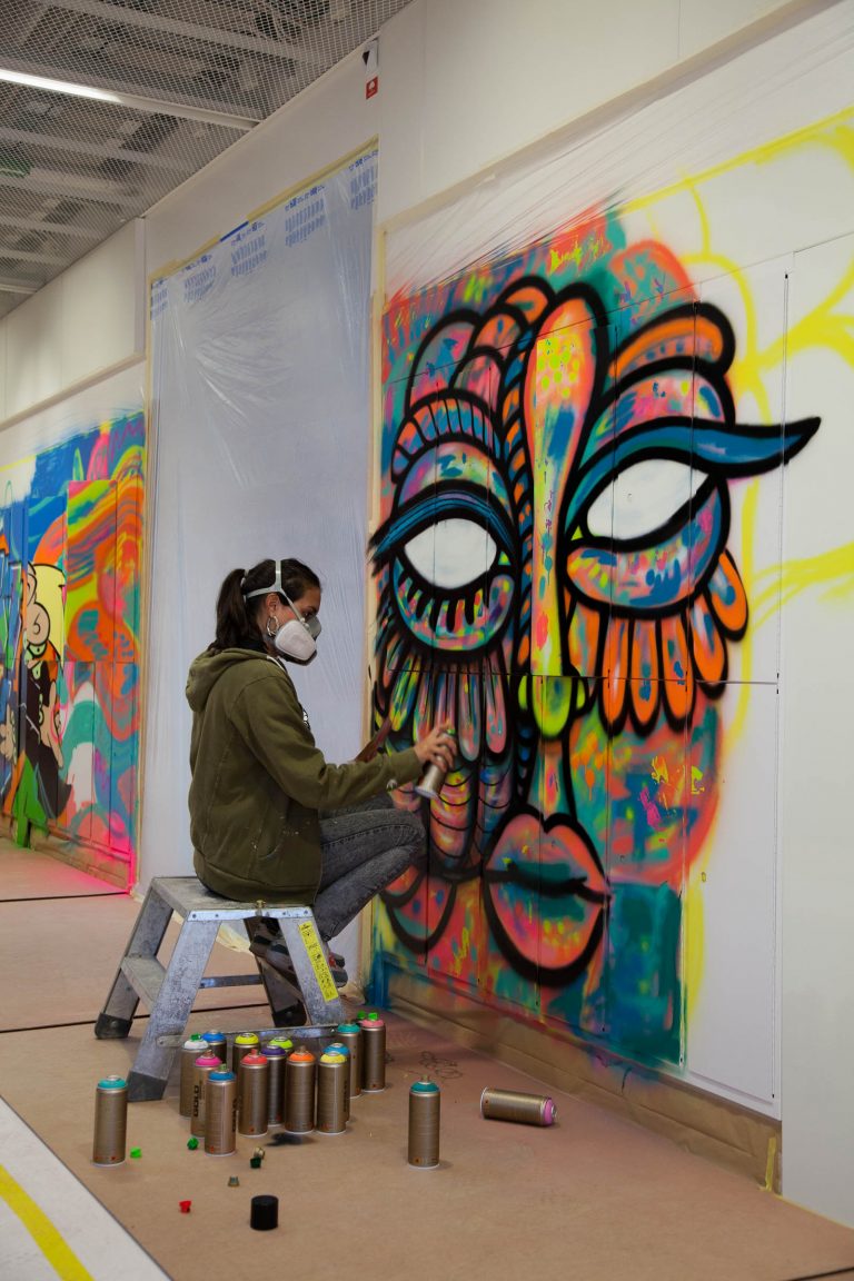 Kvinna med munskydd sitter på en pall och sprejar en graffitimålning av en färgglad fågelkvinna på väggen. På golvet sprayburkar. Pärra Andreasson, Vad Vi Vill, 2013
