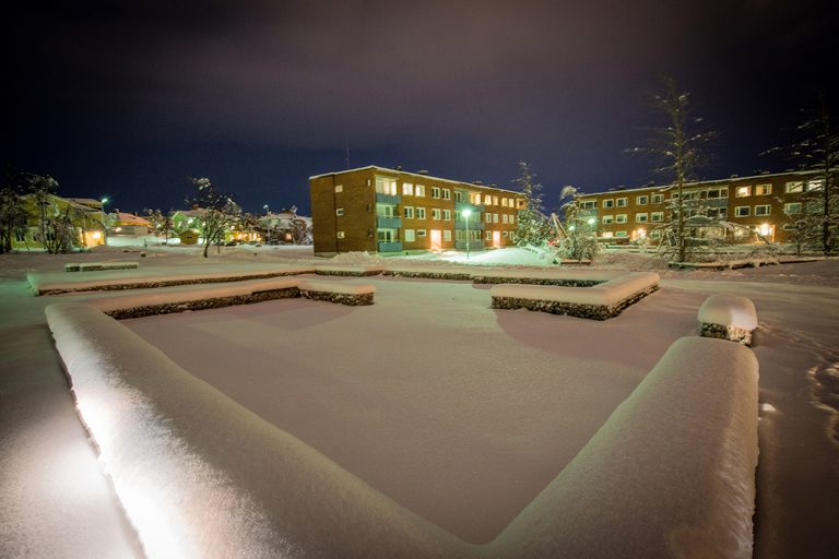 Nattbild med upplysta tegelbostadshus och trävillor i bakgrunden och låga snötäckta gabioner fyllda med rivningsmassor i förgrunden. Sofia Sundberg, Karl Tuikkanen, Ingo Vetter, Gruvstadsparken.