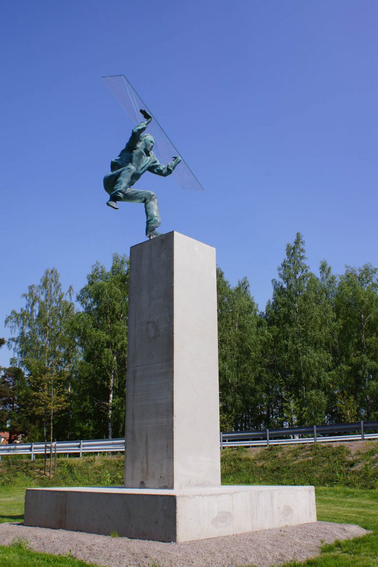 Grönärgad skulptur på en hög stensockel av en man på språng. Han har böjda knän och ena foten i luften. Heinrich Müllner, Flyer, 2014