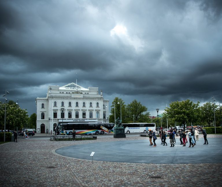 Hotfulla regnmoln över Stora Teatern i Malmö. En grupp personer i hörnet av bilden gör armrörelser. Arbetets monument Alexandra Pirici