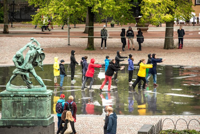 En klunga människor i regnkläder på ett torg står med armarna framsträckta. Människor tittar i bakgrunden. Arbetets monument Alexandra Pirici