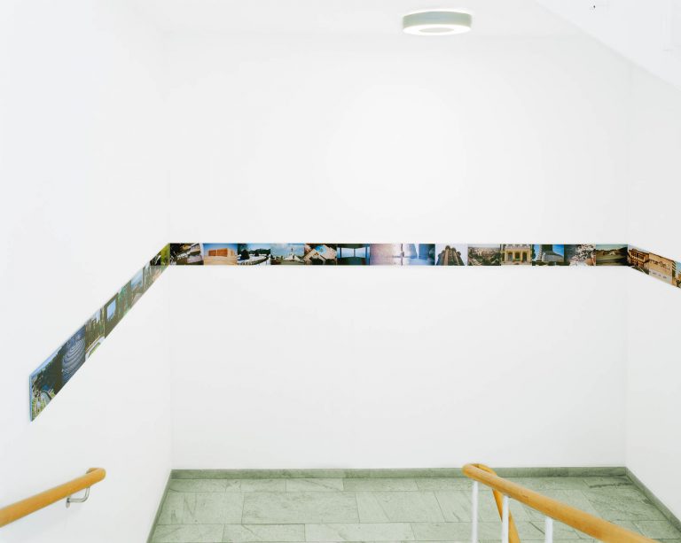Vitmålat trapphus med en smal bård av fotografier från olika platser som följer väggen. Franz Ackermann, Travelling North, (2007).