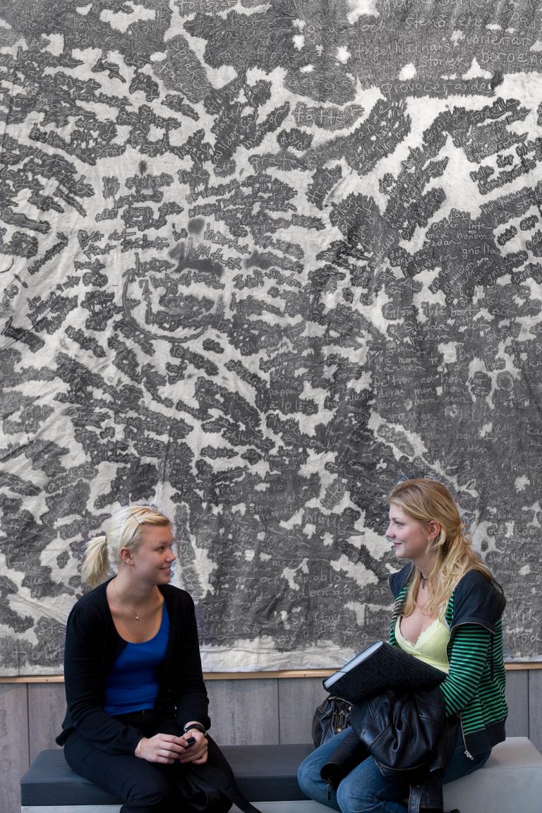 Två kvinnor sitter på en bänk framför ett stort verk i blyerts som täcker väggen bakom dem. Verket är fyllt av ord, skrivna kors och tvärs. Zsuzsanna Gilice, pssst...