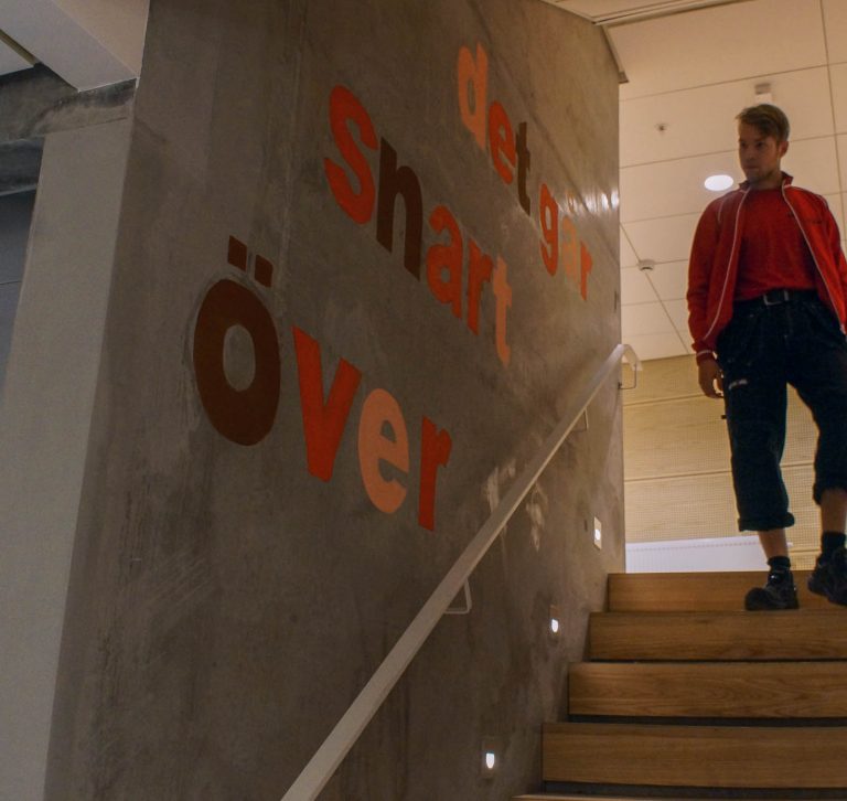 Betongvägg med ett trappräcke och olikfärgade bokstäver som bildar texten: "Det går snart över." Annika Ström, men vänta nu, 2014.