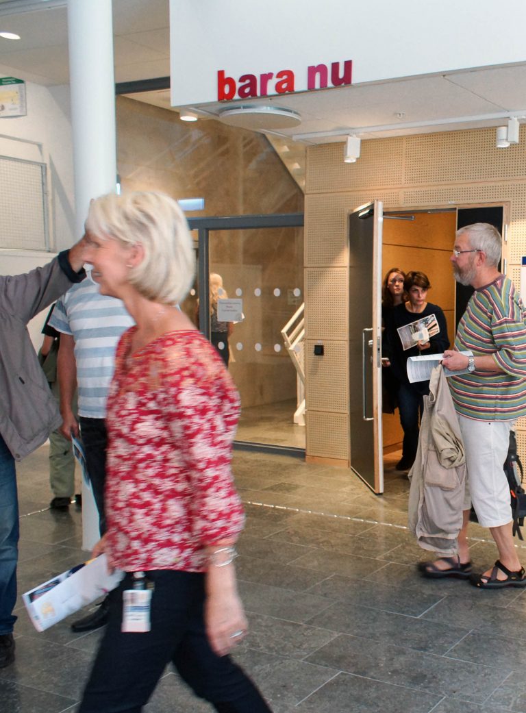 Människor i en korridor och på väg ut ur ett trapphus. På den vita väggen ovanför dörren står med röda bokstäver orden: "bara nu". Annika Ström, men vänta nu, 2014.
