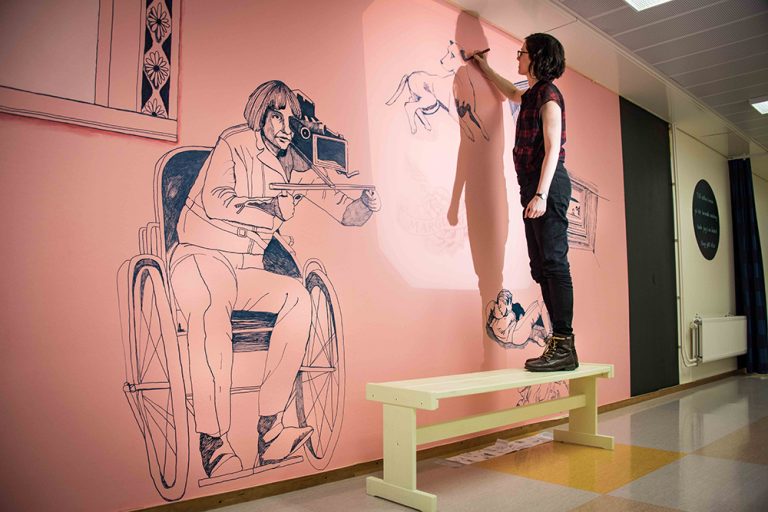 Konstnär Gerd Aurell står på en träpall och tecknar på väggen. Hon har avslutat en teckning av en man i rullstol med en märklig kikare vid ögat. Till mitten hunnen.