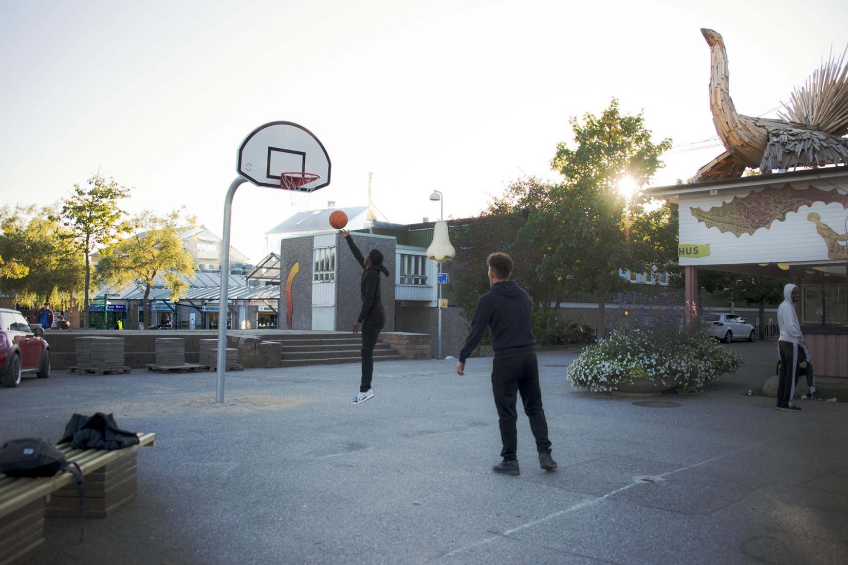 En pojke hoppar upp i luften för att kasta en basketboll i korgen, en kille står snett bakom närmare kameran. Det är en asfaltsplan utanför ungdomsgården tillika kulturhuset i Jordbro.
