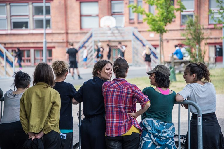 Fyra kvinnor står vid ett staket och pratar. Framför dem spelas basket på en skolgård. Myriam Lefkowitz, Walk, hands, eyes (Gamlegården)