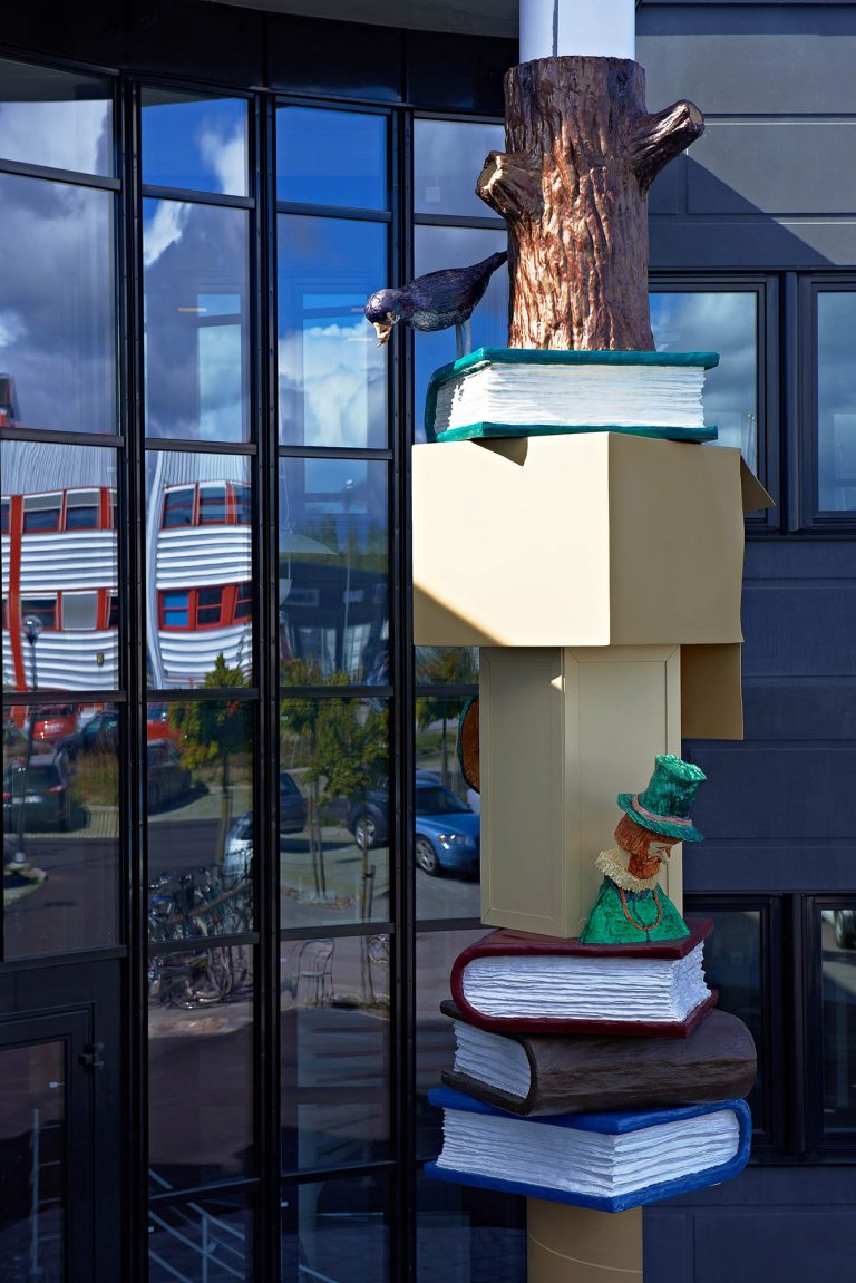 Del av pelaren med bl.a. en liten man i grön hatt och ovanför en svart fågel som kikar ner från en grön bok. Daniel Jensen, Fundamentet (2012)