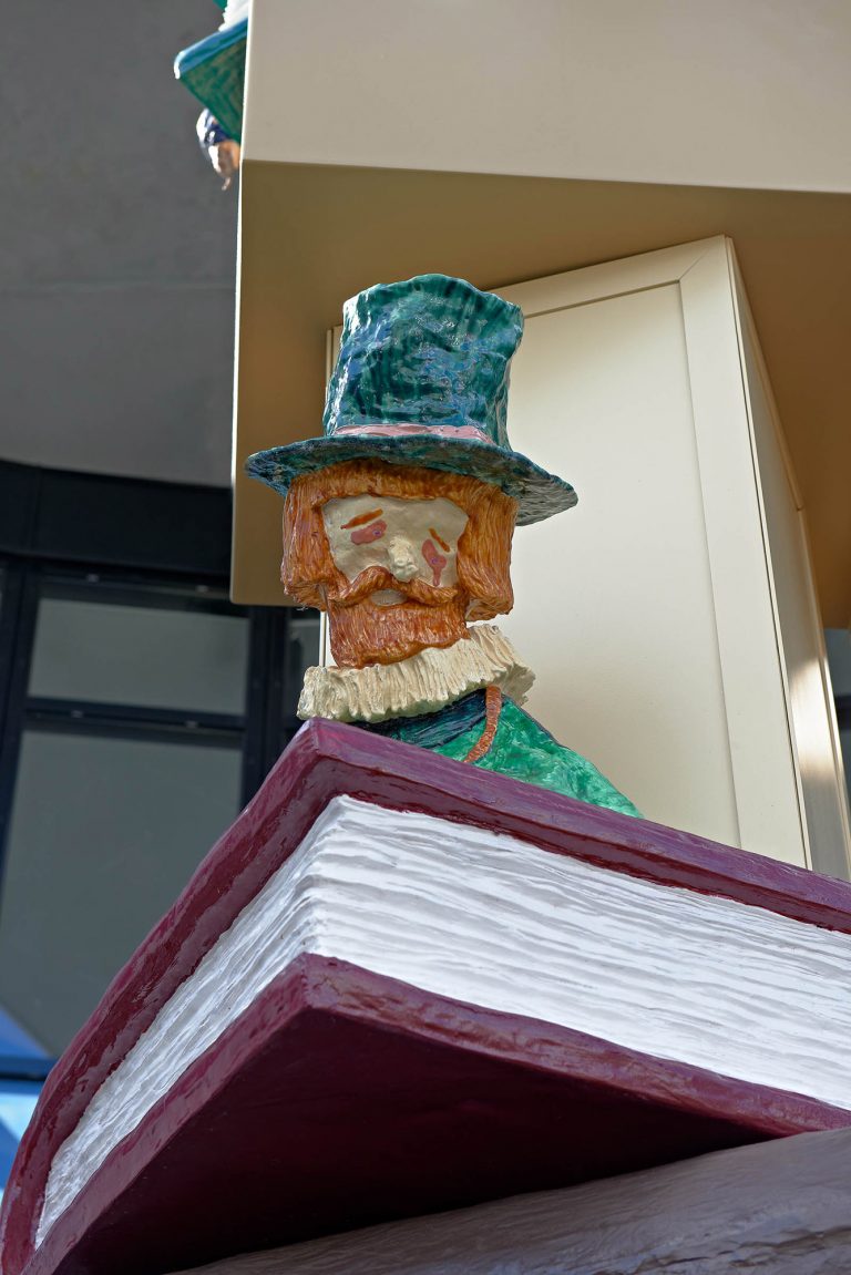En sorgsen man i grön hatt med morotsfärgat hår och pipkrage, sitter på en stor röd bok. Del av skulptural pelare. Daniel Jensen, Fundamentet (2012)