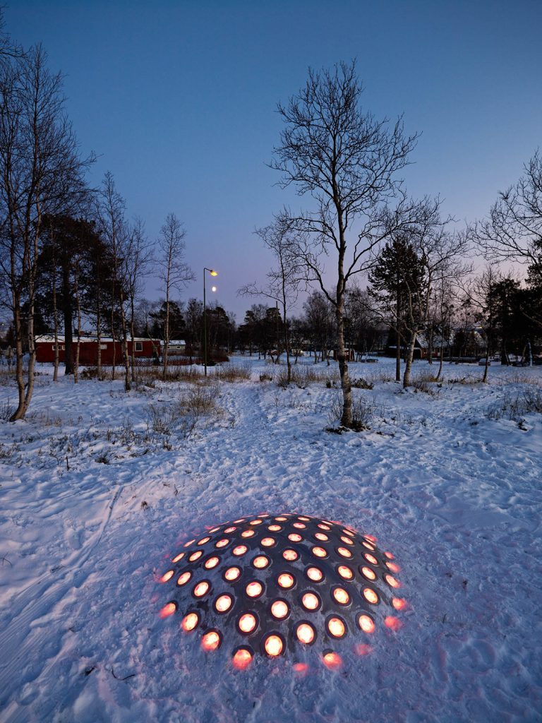 Ur den nedtrampade snön sticker stålkupolen upp vars lampor lyser orangea i halvmörkret. I bakgrunden träd och röda byggnader. Markus Lantto, Termonoder, 2009
