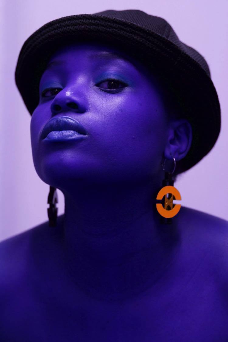En kvinna i hatt vars hud är färgad lila med orangea örhängen.