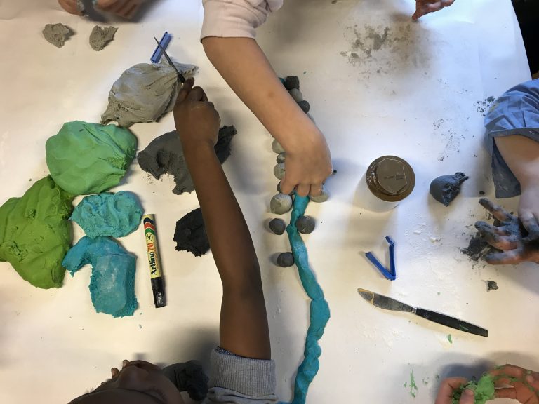 Händer arbetar med lera i olika färger och bygger landskap på ett bord.